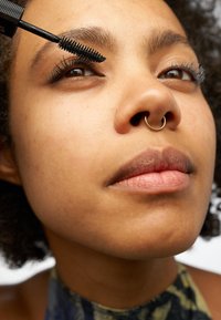 Du mascara noir est appliqué sur de longs cils noirs. Gros plan sur une peau à la texture lisse, avec un anneau de septum et une couleur de lèvres neutre.