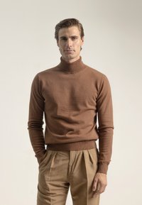 Hombre con suéter de cuello alto marrón y pantalones beige con pliegues, de pie con una mano en el bolsillo frente a un fondo claro y liso.