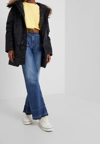 Parka nera con rifinitura in pelliccia, felpa gialla, jeans a vita alta svasati blu e sneaker bianche con lacci beige. La cintura presenta una fibbia decorativa.
