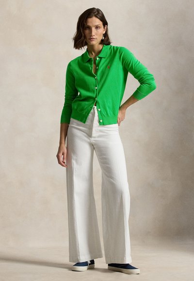 Πράσινη ζακέτα με γιακά, κλείσιμο με κουμπιά και ριμπ μανσέτες. Συνδυάζεται με λευκό παντελόνι φαρδιάς γραμμής και μπλε αθλητικά, με ουδέτερο φόντο.