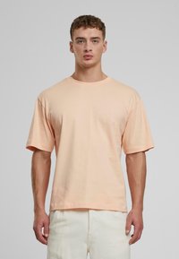 Pfirsichfarbener Baumwoll-T-Shirt mit lässiger Passform, Rundhalsausschnitt und kurzen Ärmeln, getragen mit hellen Hosen. Strukturierter Stoff, minimalistisches Design.