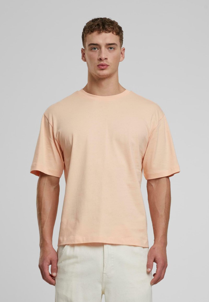 Pfirsichfarbener Baumwoll-T-Shirt mit lässiger Passform, Rundhalsausschnitt und kurzen Ärmeln, getragen mit hellen Hosen. Strukturierter Stoff, minimalistisches Design.