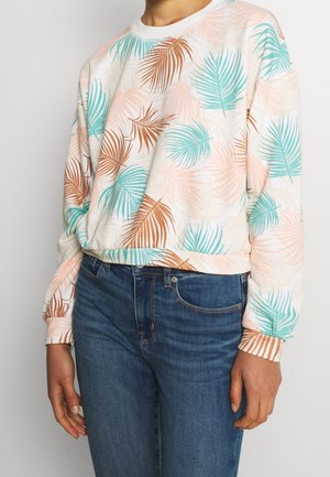 Personne portant un jean bleu et une chemise à manches longues blanche avec des imprimés de feuilles de palmier en pastel turquoise, pêche et marron.