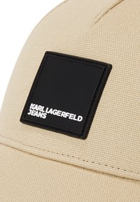 Gorra beige hecha de tela texturizada que presenta un parche cuadrado negro con el texto blanco "KARL LAGERFELD JEANS", ubicado en la parte frontal.