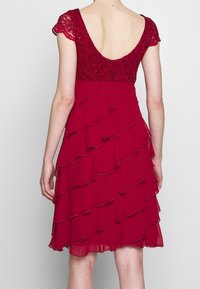 Czerwona sukienka z koronkowym gorsetem i rękawami typu cap sleeve, z warstwowym designem falbanek. Tył ma głęboki dekolt i gładką teksturę.