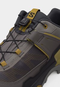 Γκρι και κίτρινο αθλητικό παπούτσι με ύφασμα από δίχτυ, ρυθμιζόμενο λουρί, ελαστικά κορδόνια και μαύρο πλαστικό κλείδωμα κορδονιών σε λευκό φόντο.