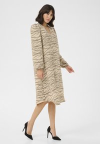 Beige klänning med vågiga svarta linjer, V-ringning, långa puffärmar och mittlång fåll. Buren med svarta högklackade skor. Slät textur.