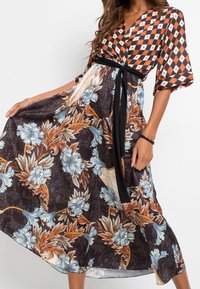Donna che indossa un vestito a fantasie miste con una parte superiore a scacchi arancione e nera, una gonna a fiori e una cintura nera, mentre tiene la gonna sollevata.