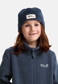 Marineblaue Fleece-Mütze mit Umschlag und Logopatch; weiche Textur und enge Passform. Getragen mit einer passenden marineblauen Fleece-Jacke mit Reißverschluss.