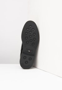 Black rubber shoe sole featuring a textured pattern with circular grips and a logo label. Flat design with a rounded toe and sleek edges.