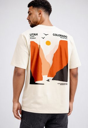 Mann trägt cremefarbenes T-Shirt mit Grafik von roten Felsenschluchten, einem Kajakfahrer, Sonne, Vögeln und Text, der Utah, Colorado und Glen Canyon erwähnt.