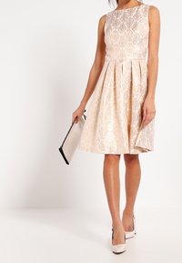 Krajková koktejlová šaty s přiléhavým korzetem a rozšířenou sukní v jemné béžové barvě. Bezramenný design s propracovanými květinovými vzory. V kombinaci s psaníčkem.