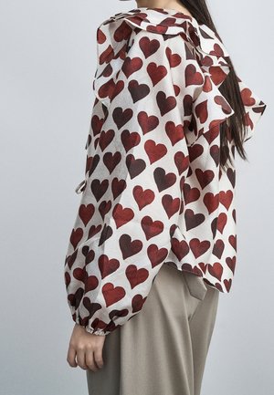 Femme portant un chemisier blanc avec des motifs de cœurs rouges et des volants aux épaules, associé à un pantalon large beige.