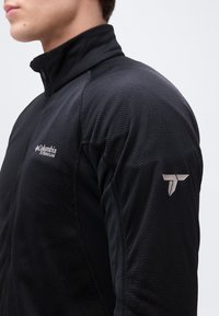 Κοντινό πλάνο ενός ατόμου που φοράει μαύρο μπουφάν Columbia Titanium με φερμουάρ, υφασμάτινη υφή και λογότυπο στο στήθος και το μανίκι.