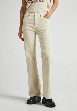 Persoon draagt beige rechte broek met hoge taille, zwarte stevige schoenen en een crème T-shirt met rode tekst dat in de broek is gestopt.