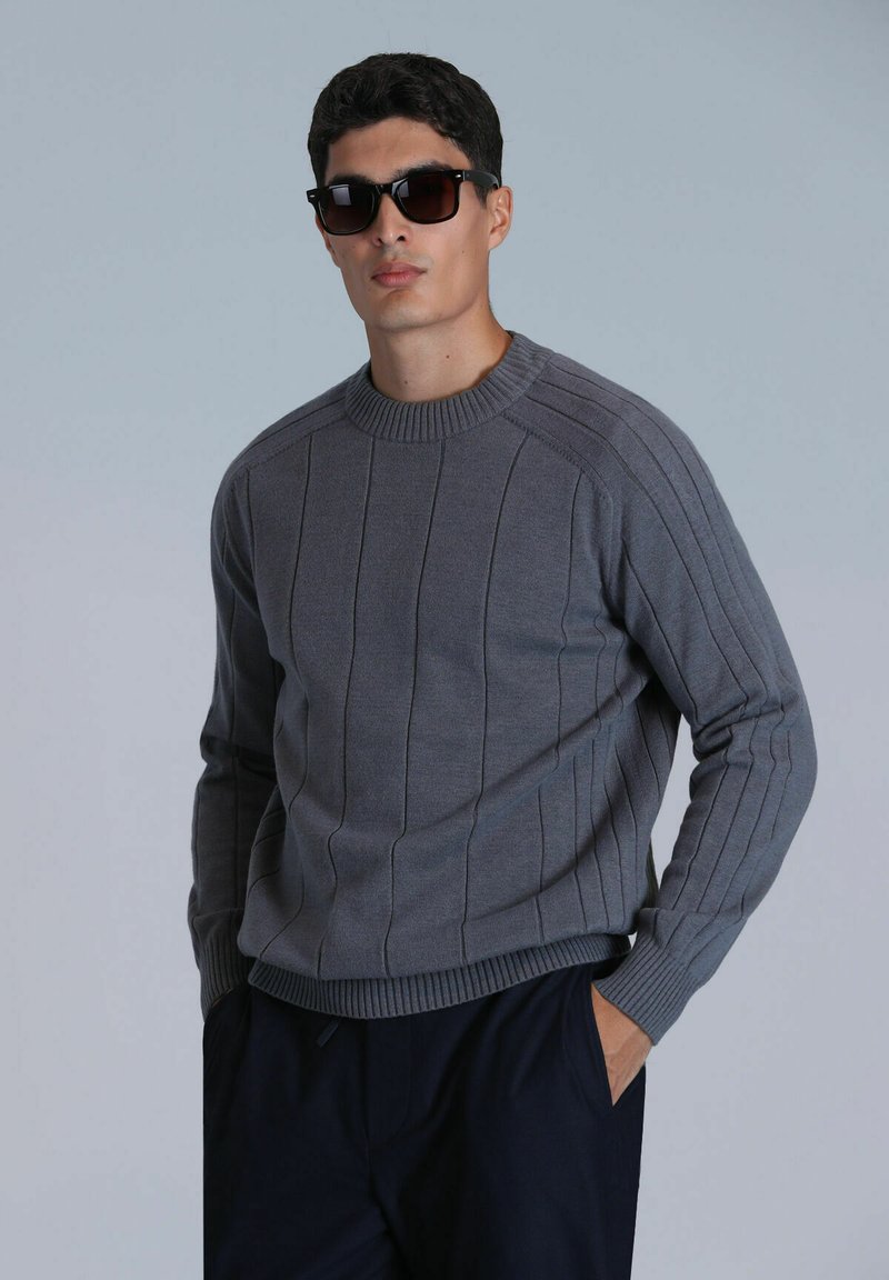 Pull gris à côtes avec un col rond, présentant des rayures verticales et des détails texturés, associé à des lunettes de soleil foncées.