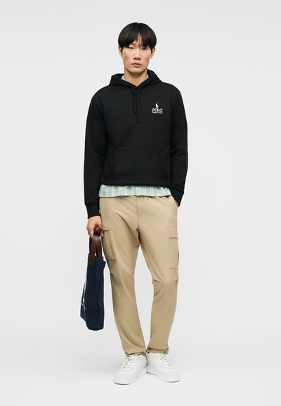 Νέος άνδρας που φοράει μαύρο φούτερ Polo, μπεζ παντελόνι cargo, λευκά αθλητικά παπούτσια, κρατώντας σκούρα μπλε τσάντα tote με καφέ χερούλια, στέκεται σε απλό φόντο.