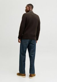 Brauner gestrickter Pullover mit hohem Kragen, kombiniert mit blauen Jeans. Das Model trägt braune Schuhe. Das Bild zeigt die Rückansicht.