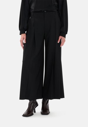 Pantaloni neri a gamba larga con pieghe, vita alta e chiusura con bottone. Il tessuto appare liscio e leggero, abbinato a stivali alla caviglia marroni.