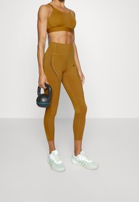Sujetador deportivo de bronce y leggings de cintura alta, ambos con una textura suave y costuras en contraste. Pesas rusas negras en la mano.