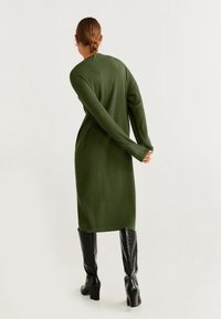 Grünes geripptes Strickkleid mit langen Ärmeln, knielang, kombiniert mit schwarzen glänzenden Overkneestiefeln, von hinten betrachtet. Glatte Textur.