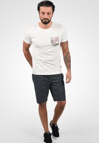 T-shirt léger pour homme de couleur claire avec une poche à motifs, associé à un short foncé. Le style global est décontracté, avec un col rond.
