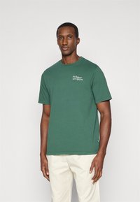 Πράσινη βαμβακερή μπλούζα t-shirt με κοντό μανίκι, στρογγυλή λαιμόκοψη, με λευκό λογότυπο και κείμενο στην αριστερή πλευρά του στήθους, απλός σχεδιασμός και κανονική εφαρμογή.