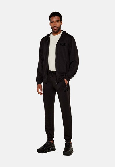 Sudadera negra con cremallera y joggers a juego, hechos de una tela suave, con detalles de logo. El modelo lleva una camisa clara y zapatillas deportivas negras.