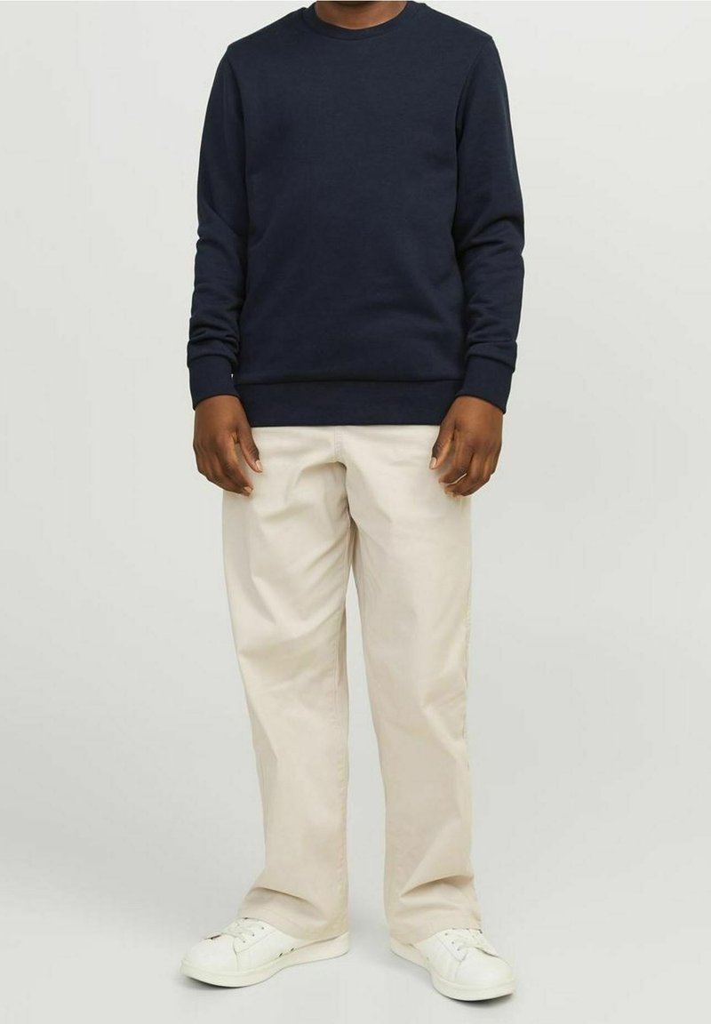 Sweat-shirt bleu marine avec col rond et poignets côtelés, assorti à un pantalon beige clair et des baskets blanches, debout devant un fond neutre.