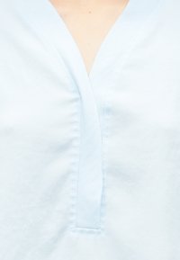 Helesinine pluus V-kaelusega, millel on õrn tekstuur ja dekoratiivne õmblus kaeluse ning keha ümber.