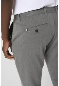Gros plan d'une personne portant un pantalon gris sur mesure avec une poche arrière boutonnée et un petit détail en tissu rayé sur le bord de la poche.