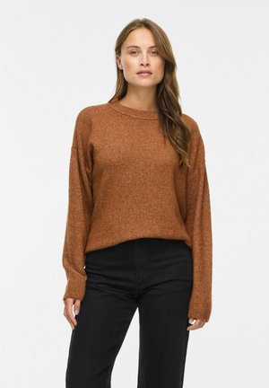 Kvinde med lysebrunt hår iført en rustfarvet strikket sweater og sorte bukser, stående mod en ensfarvet hvid baggrund.