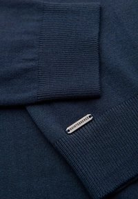 Merinokangas tummansinisessä neuleessa; joustavat hihansuut; pieni, hopeinen "Quintessence" -tikkamerkki helman kohdalla.