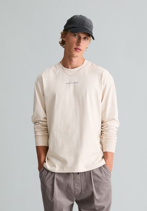 Jonge man die een beige overhemd met lange mouwen, grijze broek, zwarte pet en een ketting draagt, staand met handen in de zakken tegen een effen achtergrond.