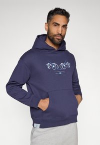 Sudadera azul marino hecha de una mezcla de algodón, con un bolsillo frontal y un diseño gráfico en colores más claros. Textura suave y corte casual.