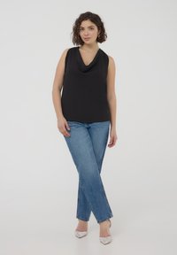 Top nero senza maniche con scollatura drappeggiata, abbinato a jeans blu chiaro a gamba diritta e tacchi a punta argentati su uno sfondo semplice.