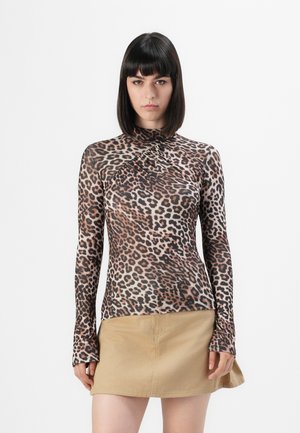 Vrouw in een lang mouw leopardprint top met een hoge hals en een beige mini-jurk, staande tegen een vlakke witte achtergrond.
