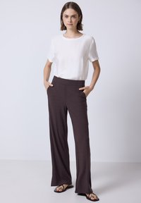 Bruine geribbelde wijde broek met een elastische tailleband, gecombineerd met een eenvoudige witte top met korte mouwen. Het model staat met de handen in de zakken.