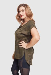 Grønn camo kortermet topp i myk stoff, med rund utringning og sideslisser, sammen med gjennomsiktige svarte leggings.
