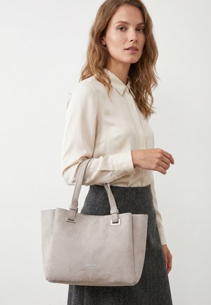 Woman wearing cream blouse and gray skirt carrying light gray suede tote bag on shoulder against plain background.