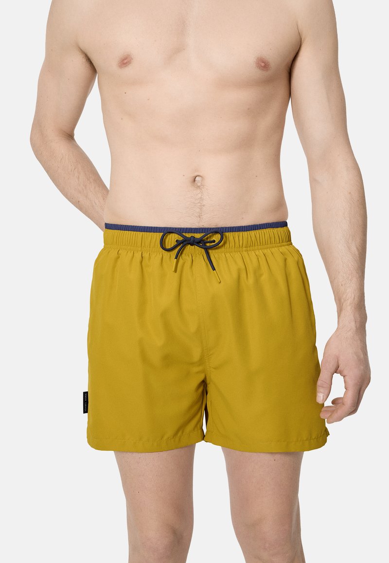 Hombre con pantalones cortos de baño amarillos mostaza con cintura elástica azul marino y cordón negro sobre un fondo claro y liso.