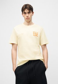 Νεαρός με κοντά καστανά μαλλιά φοράει μπεζ μπλουζάκι Nike με πορτοκαλί γράμματα και μαύρο παντελόνι, στέκεται μπροστά σε απλό φωτισμένο φόντο.