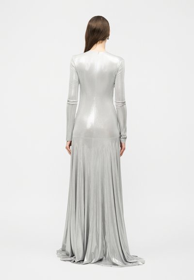 Γυναίκα με μακριά μαλλιά που φοράει εφαρμοστή ασημί μεταλλική τουαλέτα με μακριά μανίκια και ρέουσα πλισέ φούστα, εμφανιζόμενη από πίσω.