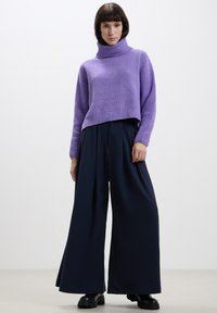 Maglione a collo alto a coste viola abbinato a pantaloni larghi blu navy. La modella indossa stivali neri chunky; sfondo minimalista.