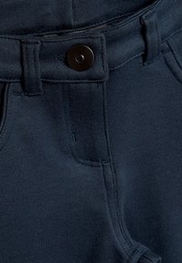 Detailný záber na tmavomodrý pás nohavíc, zobrazujúci čierny gombík, slučky na opasok a viditeľné švy.