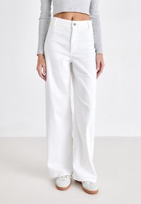 Witte wijde denimbroek met hoge taille, knoopsluiting aan de voorkant en een soepele textuur. Gecombineerd met lichte sneakers.