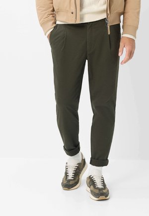 Mand iført mørke olivenfarvede tapered bukser med opbrettede manchetter, hvide sokker, sneakers med camouflage mønster, beige jakke og cremefarvet sweater med hænderne i lommerne.