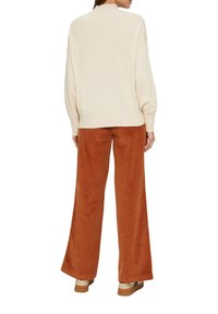 Pull en maille beige à col montant, associé à un pantalon large en velours côtelé couleur rouille. Les chaussures présentent un design superposé et des tons neutres.