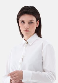 Camicia bianca con una placca arricciata e bottone decorativo. Maniche lunghe con polsini abbottonati, tessuto liscio e design classico.