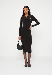 Fekete, hosszú ujjú polóruha gallérral és gombos részlettel, karcsúsított szabással. Fekete ívelt kézitáskával és hegyes orrú magassarkú cipővel kombinálva.